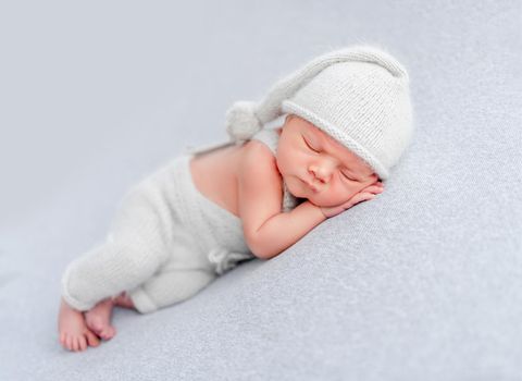 Newborn baby boy wearing knitted pants and hat sleeping and holding tiny hands under his cheeks. Adorable infant child napping in studio