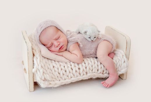 Adorable newborn baby boy sleeping on his tummy and little fluffy kitten lying on his back. Cute infant kid wearing knitted costume and hat napping with small cat kity during studio photoshoot