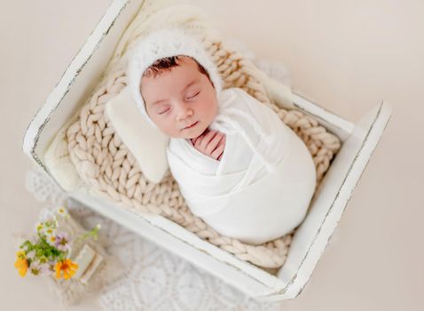 Little beautiful newborn baby girl swaadled in fabric and wearing knitted hat sleeping in tiny designed bed during studio photoshoot. Cute infant child kid napping resting