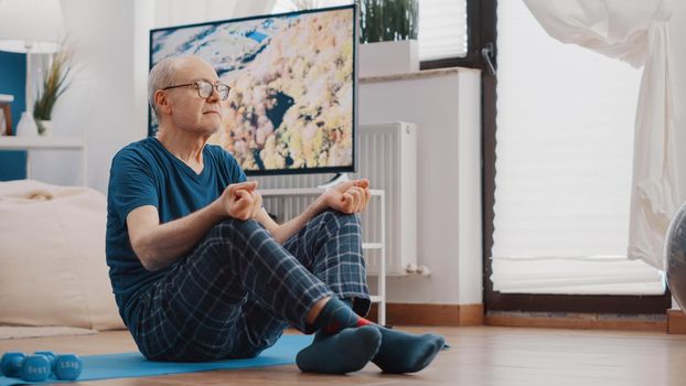 Calm person sitting in lotus position to meditate on mat. Senior man in yoga pose meditating for wellness and relaxation at home. Retired person with eyes closed doing meditation.