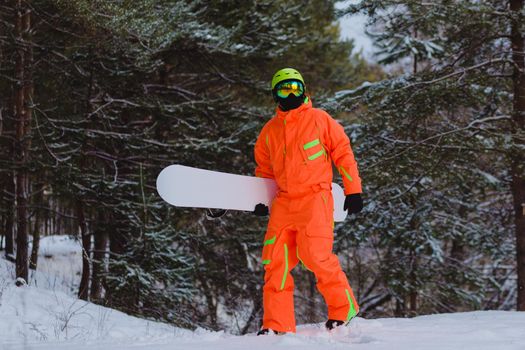 Snowboarder dressed in orange suit walking through the forest