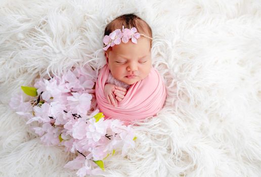 Swaddled beautiful newborn baby girl with flower wreath on her head sleeping in white fur. Cute female infant child photoshoot in studio. Adorable kid napping