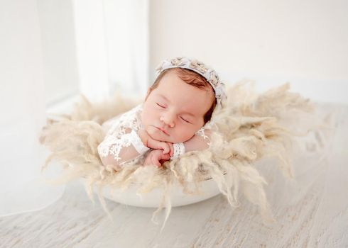 Light portrait of little beautiful newborn baby girl wearing wreath sleeping in basin with fur decoration during studio photoshoot. Cute infant child napping holding hands ander cheeks