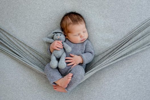 Newborn baby boy wearing knitted costume sleeping in hammock and holding toy in hands. Infant kid studio portrait with decoration