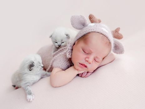 Adorable newborn baby boy sleeping on his tummy and two little fluffy kittens sitting close to him. Cute infant kid wearing knitted costume and hat napping with cats during studio photoshoot