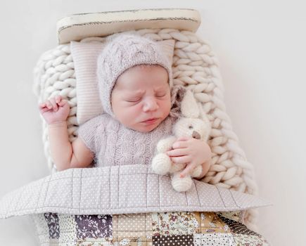 Adorable newborn baby boy sleeping in wooden stylized bed under blanket and holding toy. Cute infant kid wearing knitted costume and hat napping during studio photoshoot