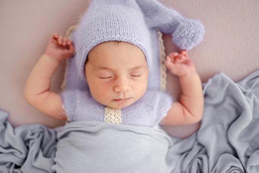 Beautiful little newborn baby girl wearing knitted costume and hat sleeping with hands up under blanket in studio. Cute infant child napping