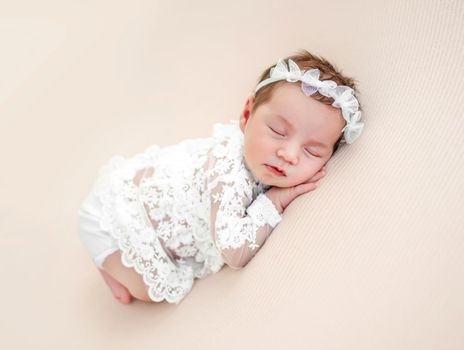 Adorable newborn baby girl wearing beautiful white costume and wreath lying on her tummy and sleeping in studio. Cute infant child napping holding hands under cheeks