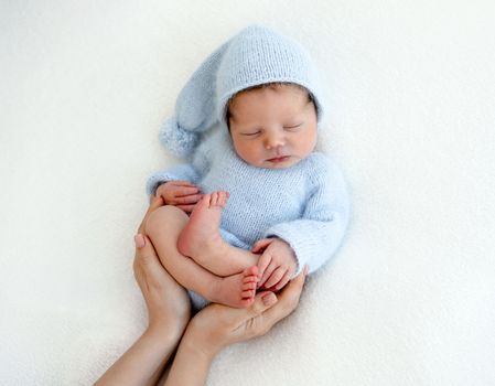 Sweet newborn baby boy sleeping wearing knitted costume and his mother hands care about him. Napping infant kid studio portrait with parent