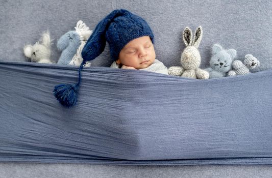 Adorable newborn baby boy wearing cute knitted hat sleeping under fabric with cute handmade toys. Infant child napping indoors