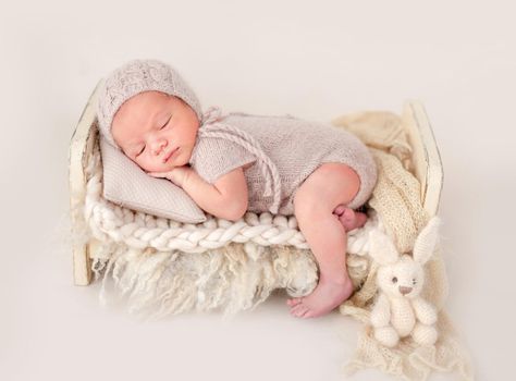 Newborn baby boy wearing knitted beige costume and hat sleeping on his tummy in small designed bed and holding tiny hands under his cheeks. Adorable infant child napping