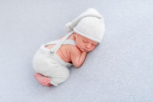Newborn baby boy wearing knitted pants and hat sleeping on his tummy and holding tiny hands under his cheeks. Adorable infant child napping in studio with light blue colors