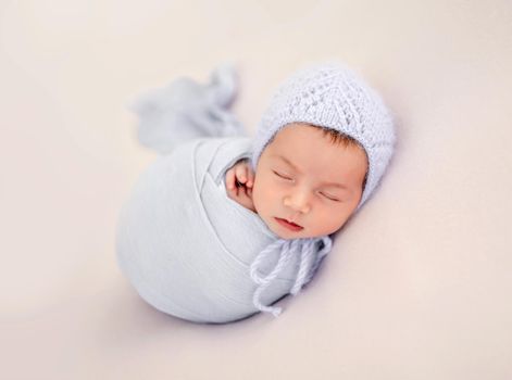 Little beautiful newborn baby girl swaadled in fabric and wearing knitted hat sleeping during studio photoshoot. Cute infant child kid napping resting