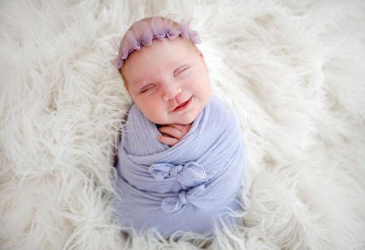 Newborn baby girl swaddled in light blue fabric sleeping and smiling. Little cute infant child napping lying on white fur. Adorable kid during studio photoshoot