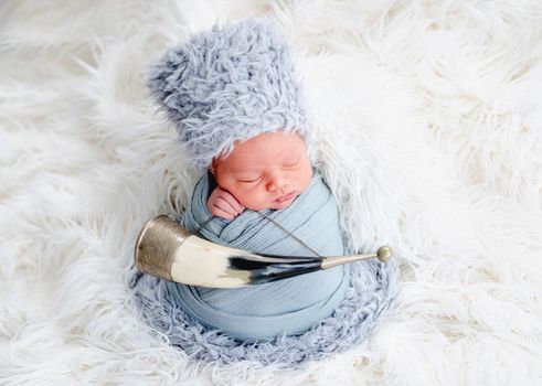 Portrait of newborn baby boy swaddled in light blue fabric and wearing furry caucasian hat sleeping in studio. Adorable infant child napping with traditional horn