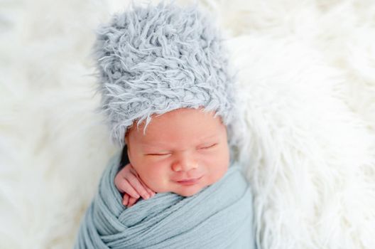 Portrait of newborn baby boy swaddled in light blue fabric and wearing furry hat sleeping in studio. Adorable infant child napping