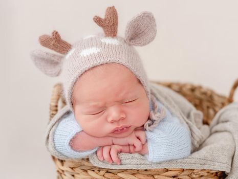 Portrait of newborn baby boy wearing knitted hat with deer horns sleeping on his tummy and holding tiny hands under his cheeks. Adorable infant child napping