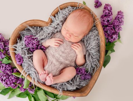 Newborn baby boy wearing knitted beige costume lying in wooden heart shape bed with purple flowers decoration and sleeping. Adorable infant child napping