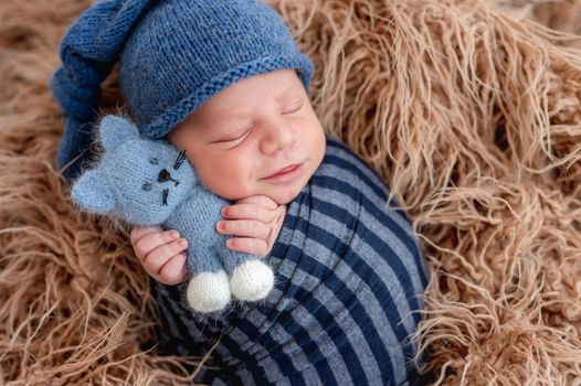 Adorable newborn baby boy swaddled in toby fabric holding in his tiny hands knitted toy and smiling during sleeping. Cute infant kid wearing hat napping in fur during studio photoshoot.