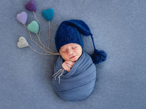 Newborn baby boy swaddled in blue fabric and wearing hat sleeping and holding knitted toy hearts. Photo composition with infant kid napping on blue backgroung