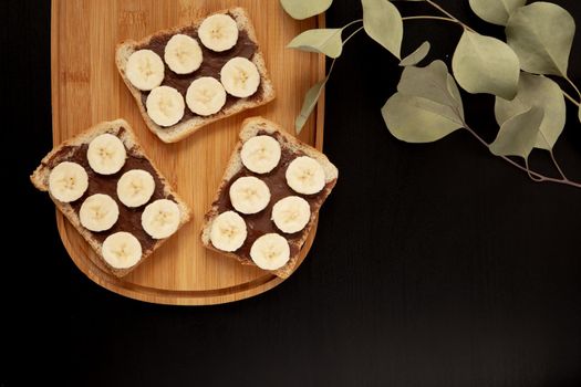 Three banana white bread toasts spread with chocolate butter that lie on a cutting board with a sprig of leaves on a dark background. top view with area for text