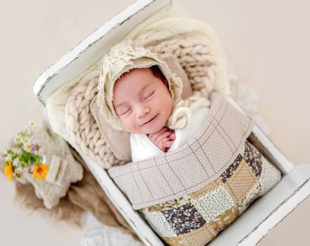 Little beautiful newborn baby girl wearing hat sleeping under handmade blanket in tiny designed bed and smiling during studio photoshoot. Cute infant child kid napping resting