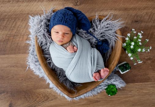 Adorable newborn baby boy swaddled in fabric and wearing cute knitted hat falling asleep during studio photoshoot in wooden heart shape bed. Infant child napping