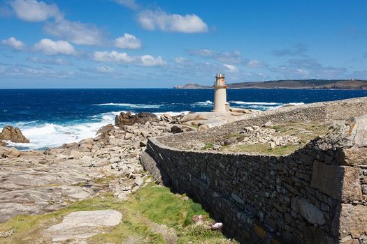 View on a lighthouse and ancient wall made of stones near the ocean with blue skies and white clouds. The scene from Camino de Santiago, tourist path in Spain.