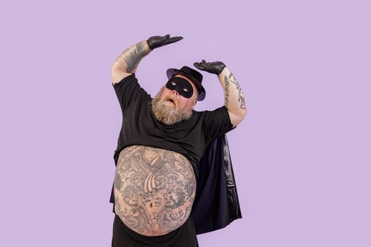 Doubting mature obese gentleman wearing hero costume with cape and mask covers himself from upper danger by hands on purple background in studio