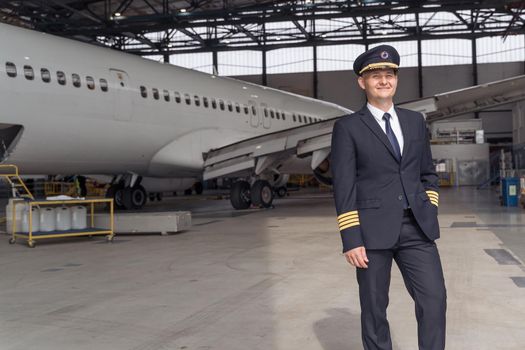 Happy pilot in uniform ready for flight while standing in front of big passenger airplane in airport