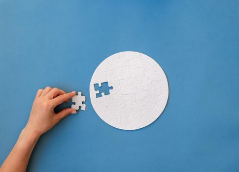 Hand putting last piece in white round shaped jigsaw puzzle on blue background