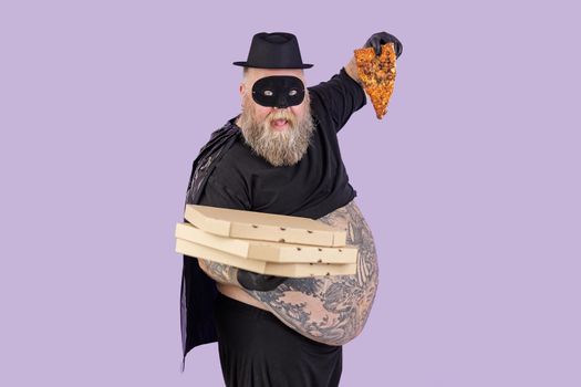 Funny plump man in hero suit with large bare abdomen holds boxes stack and slice of tasty pizza standing on purple background in studio