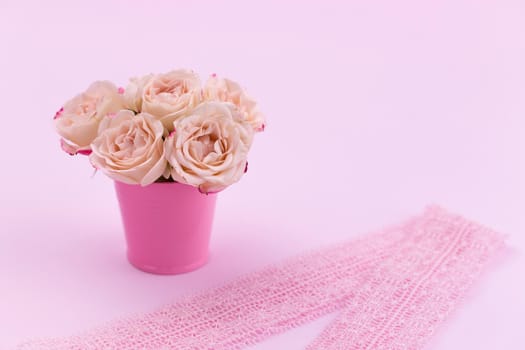 A bouquet of beautiful roses stands in a small bucket on a lace ribbon on a pink background with space for text