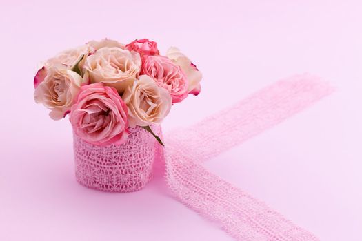 A bouquet of beautiful roses stands in a small bucket on a lace ribbon on a pink background with space for text