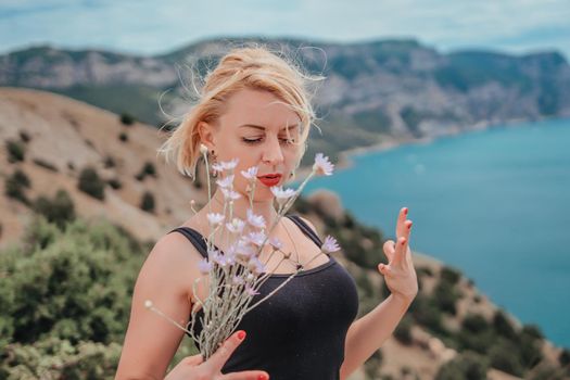 blonde girl with scarlet lipstick and red manicure in a black T-shirt with a bouquet of wildflowers on a background of sea and mountains. she is happy and enjoying wonderful breathtaking landscape.
