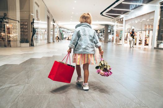 Small cute fashion child is carrying shopping bag and flowers inside the Shopping Mall after black Friday sale. View from behind. Toddler is happy and feels she is a shopoholic.