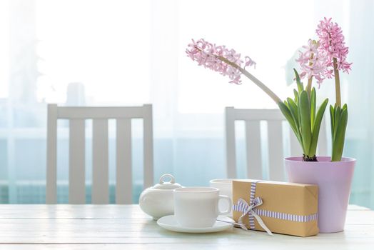 Present gift box with white cups of tea and pink flowers on the white table near window. Lovely spring holiday mood