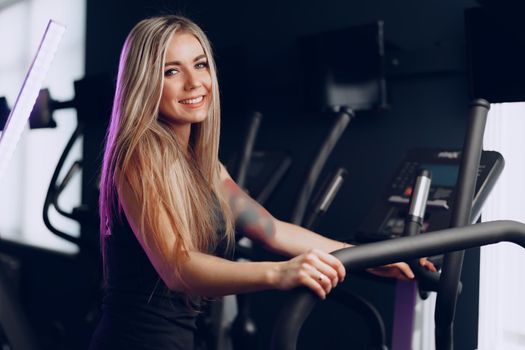 Attractive young blonde woman exercising on cardio training apparatus in dark gym