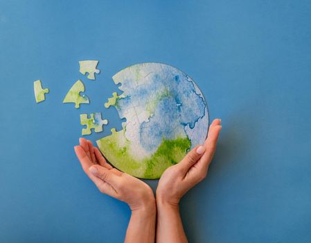 Hands holding unfinished jigsaw puzzle in shape of globe on blue background. Protecting the planet concept