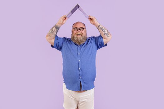 Joyful mature man with overweight in tight blue shirt with glasses holds laptop above head as roof on purple background in studio