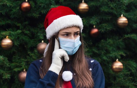 Choosing a gift. Close up Portrait of woman wearing a santa claus hat and medical mask with emotion. Against the background of a Christmas tree. Coronavirus pandemic