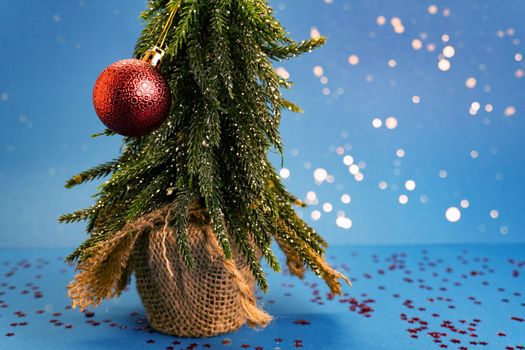 Green tree with small red fragile toy is on blue table with red stars and blue background behind the tree. New year and christmas concept, bokeh. Copyspace for the text, good for banner.