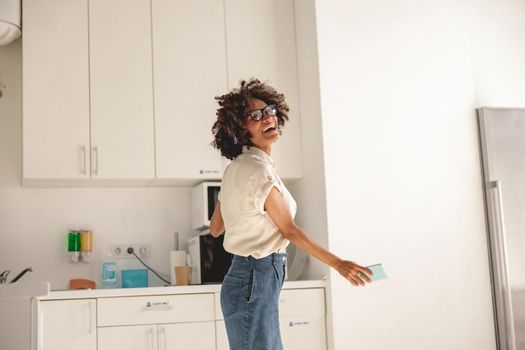 Pretty young lady wearing wireless headphones and dancing in the kitchen while holding the smartphone