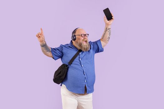 Cheerful bearded plus size man with headphones and crossbody bag enjoys music on mobile phone posing on purple background in studio