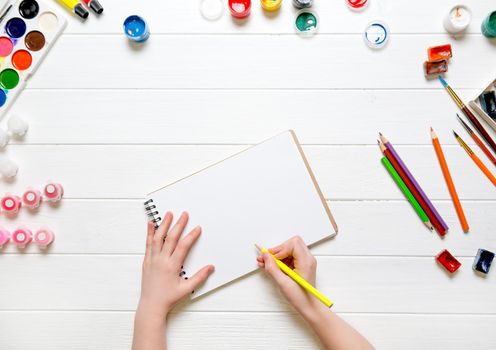 Top view of girl's hand with pencil over white blank paper and colorful pencils and watercolors on white wooden table background. Copy space