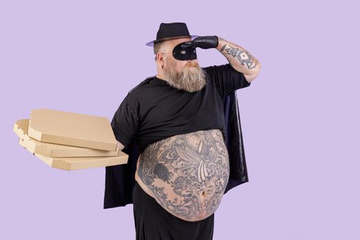 Funny man with overweight in hero costume with large tattooed tummy holds cardboard boxes with pizzas looking into distance on purple background in studio