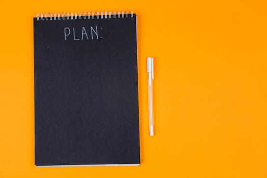 Wide view image of blank black spiral note pad and white marker with calligraphic inscription plan on yellow background