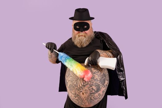 Mature man with overweight in hero costime with large bare tattooed belly sprays detergent onto fluffy brush on purple background in studio