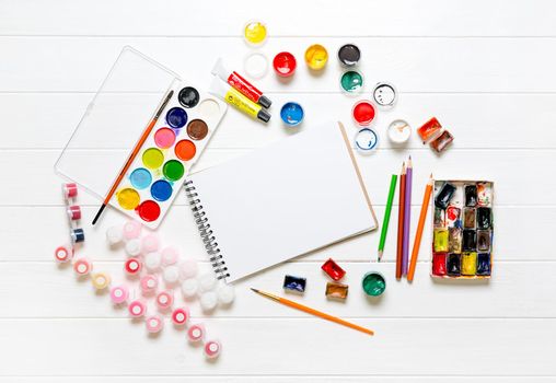 White blank notepad among drawing accessories on white wooden background. Top view of color pencils, watercolors and brushes. Copy space