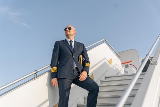 Happy adult pilot looking away while standing on stairs of aircraft. Profession concept. Copy space